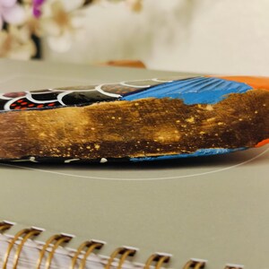 May include: Close-up of a decorative wooden object with a textured, brown surface. The object features blue, orange, and black painted details. The object is resting on a gray surface with a spiral notebook in the foreground.