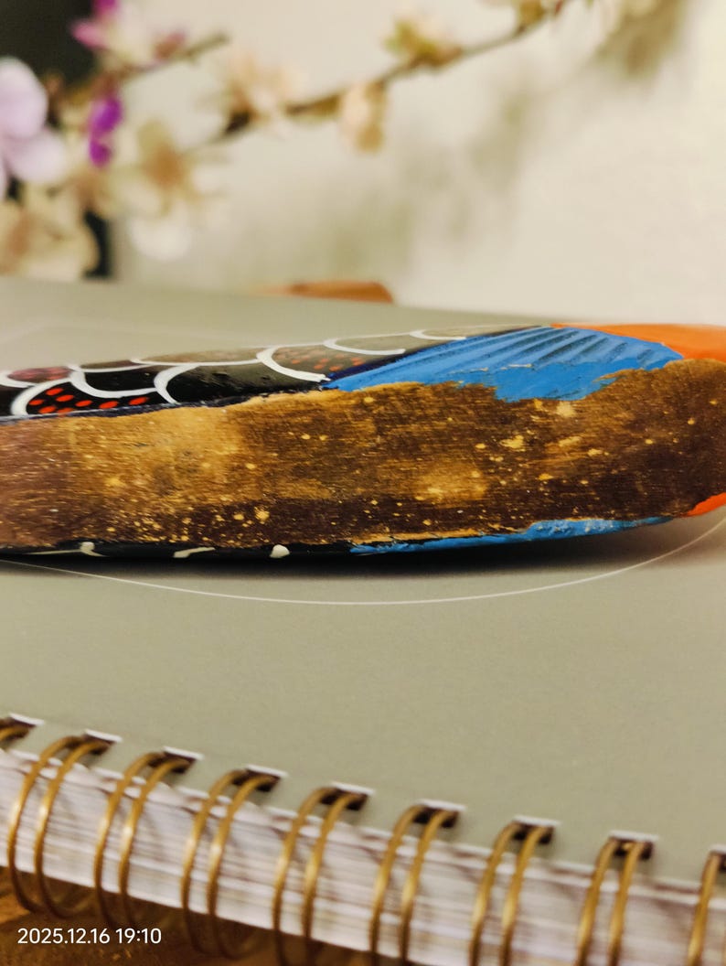 May include: Close-up of a decorative wooden object with a textured, brown surface. The object features blue, orange, and black painted details. The object is resting on a gray surface with a spiral notebook in the foreground.