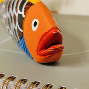 May include: A hand-painted wooden fish sculpture with an orange head, red mouth, and yellow dorsal fin. The fish has black and white patterned scales and blue accents. The sculpture is resting on a gray surface.