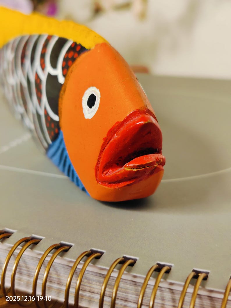 May include: A hand-painted wooden fish sculpture with an orange head, red mouth, and yellow dorsal fin. The fish has black and white patterned scales and blue accents. The sculpture is resting on a gray surface.