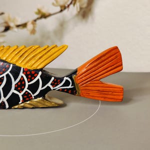 May include: A hand-painted wooden fish sculpture with a black body, white scale pattern, and red polka dots. The fins are yellow and orange. The fish is a decorative item.