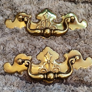 Puede incluir: Dos tiradores de cajón dorados y ornamentados. Cada tirador presenta un elemento decorativo central con un asa curvada y acentos en forma de nube a cada lado. Los tiradores son probablemente de metal y están diseñados para muebles.