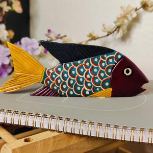 May include: A hand-painted wooden fish sculpture. The fish features a maroon head, a black dorsal fin, and a yellow tail. The body is teal with white and red scale details. The fish is resting on a gray surface.