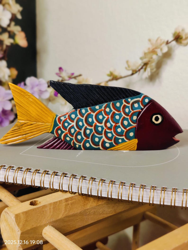 May include: A hand-painted wooden fish sculpture. The fish features a maroon head, a black dorsal fin, and a yellow tail. The body is teal with white and red scale details. The fish is resting on a gray surface.