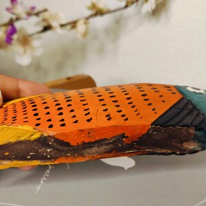 May include: Hand-painted wooden fish sculpture. The fish features orange scales with black dots, a blue head, and yellow and brown fins. The artwork is displayed against a neutral background with floral accents.