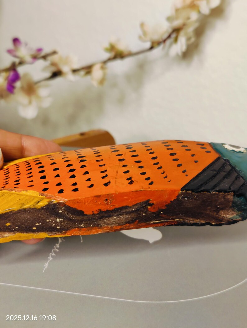May include: Hand-painted wooden fish sculpture. The fish features orange scales with black dots, a blue head, and yellow and brown fins. The artwork is displayed against a neutral background with floral accents.
