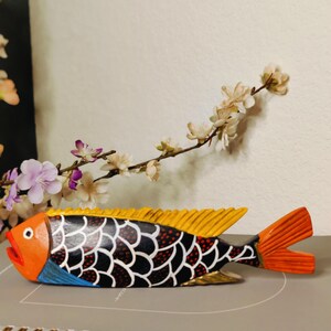 May include: A hand-painted wooden fish sculpture with orange, yellow, black, and blue details. The fish has a detailed scale pattern and a vibrant orange tail and mouth. Decorative floral accents are in the background.