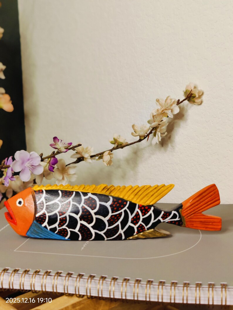 May include: A hand-painted wooden fish sculpture with orange, yellow, black, and blue details. The fish has a detailed scale pattern and a vibrant orange tail and mouth. Decorative floral accents are in the background.