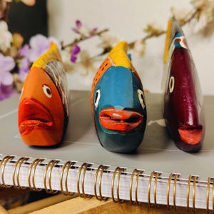 May include: Three colorful, hand-painted wooden fish figurines. The fish are orange, blue, and maroon, with yellow and red accents. They are displayed on a spiral-bound notebook, suggesting a decorative or artistic theme.