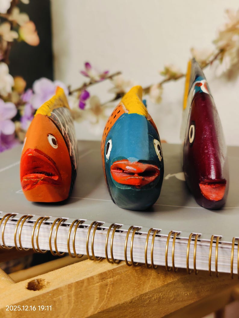 May include: Three colorful, hand-painted wooden fish figurines. The fish are orange, blue, and maroon, with yellow and red accents. They are displayed on a spiral-bound notebook, suggesting a decorative or artistic theme.