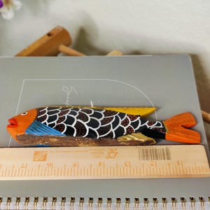 May include: A hand-carved wooden fish figurine with orange, black, and yellow details. The fish has a detailed scale pattern and a vibrant orange tail. The fish is approximately 10 inches long.