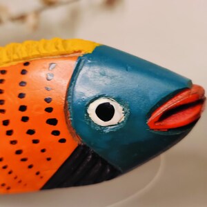 May include: A colorful, hand-painted wooden fish sculpture. The fish features a blue head, orange body with black dots, and red lips. The top fin is yellow. The sculpture is a decorative item.