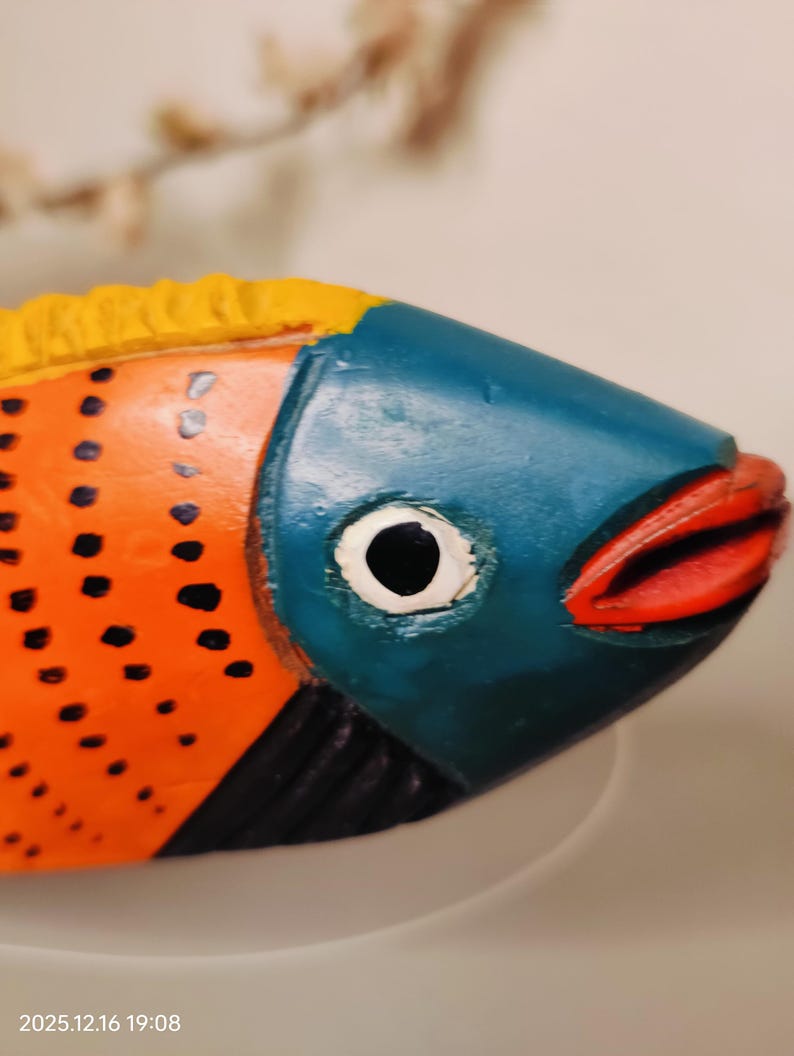 May include: A colorful, hand-painted wooden fish sculpture. The fish features a blue head, orange body with black dots, and red lips. The top fin is yellow. The sculpture is a decorative item.