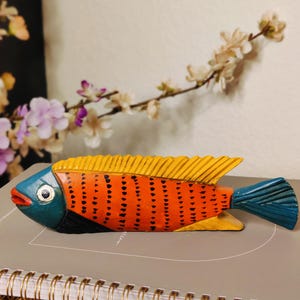 May include: A hand-carved wooden fish sculpture. The fish features a vibrant color scheme with an orange body, yellow fins, and a blue head and tail. The body is decorated with black dots. The sculpture is approximately 10 inches long.