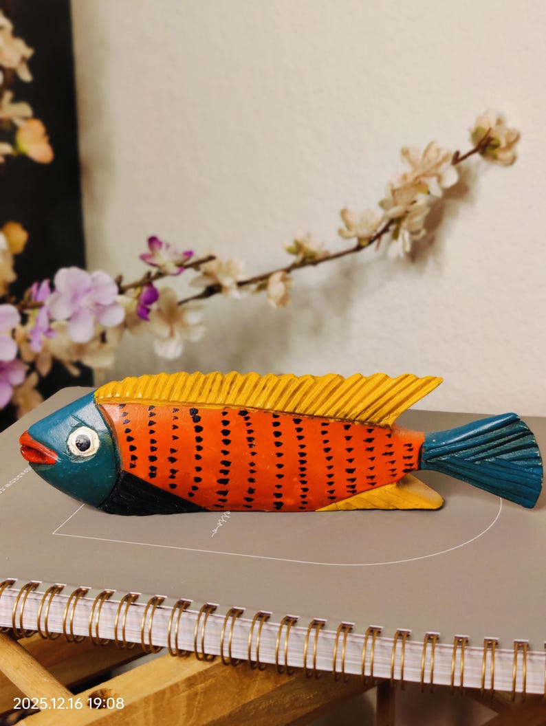 May include: A hand-carved wooden fish sculpture. The fish features a vibrant color scheme with an orange body, yellow fins, and a blue head and tail. The body is decorated with black dots. The sculpture is approximately 10 inches long.