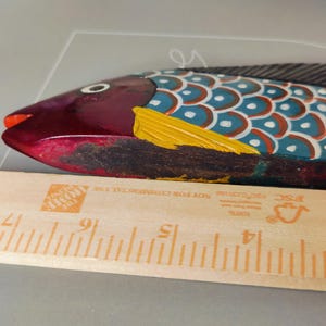 May include: A hand-painted wooden fish sculpture with a vibrant color scheme. The fish features a maroon head, a blue and white body, and a yellow fin. A wooden ruler is placed next to the fish. The fish measures approximately 10 inches in length.