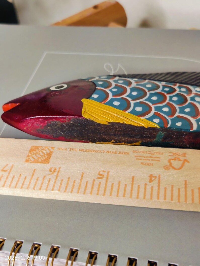 May include: A hand-painted wooden fish sculpture with a vibrant color scheme. The fish features a maroon head, a blue and white body, and a yellow fin. A wooden ruler is placed next to the fish. The fish measures approximately 10 inches in length.