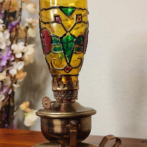 Könnte beinhalten: Eine antike Öllampe mit dekorativem Glasschirm. Der Schirm ist gelb mit farbenfrohen geometrischen Designs in Rot, Grün und Lila. Die Lampe hat einen Messingfuß und einen Griff.