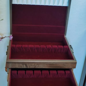 May include: Wooden jewelry box with a dark brown exterior and a burgundy velvet interior. The top section has a white padded lining and the box has two pull-out drawers with velvet compartments for storage.