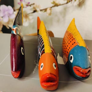 May include: Three hand-painted wooden fish sculptures. The fish have vibrant colors, including orange, blue, and red. Each fish has a unique design, with details like scales and fins. The sculptures are decorative and add a touch of whimsy.