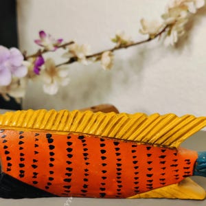 May include: A hand-carved wooden fish sculpture. The fish features an orange body with black dot patterns, yellow fins, and a blue tail. The sculpture is set against a blurred background with white and purple flowers.