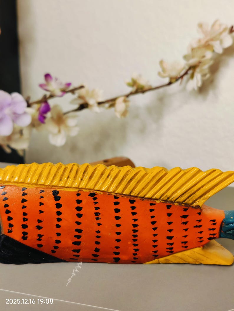 May include: A hand-carved wooden fish sculpture. The fish features an orange body with black dot patterns, yellow fins, and a blue tail. The sculpture is set against a blurred background with white and purple flowers.