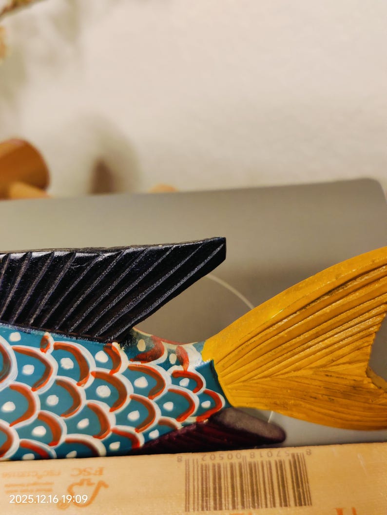 May include: A colorful wooden fish sculpture with a black dorsal fin and a yellow tail fin. The body is painted with blue, red, and white patterns. The sculpture is a decorative item.