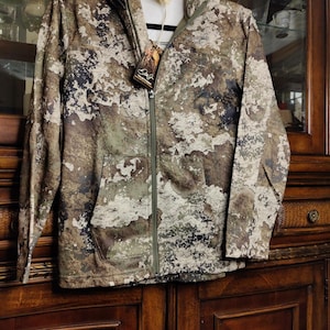 Könnte beinhalten: Eine Tarn-Kapuzenjacke mit durchgehendem Reißverschluss und einer Vordertasche. Die Jacke hat ein Muster aus Braun, Grün und Weiß und hängt an einem schwarzen Kleiderbügel. Ein Etikett ist am Reißverschluss befestigt.