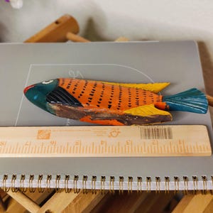 May include: A hand-painted wooden fish figurine. The fish has an orange body with black markings, yellow fins, and a blue tail. The head is blue and red. A wooden ruler is next to the fish. The fish is approximately 10 inches long.