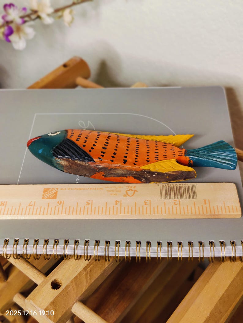 May include: A hand-painted wooden fish figurine. The fish has an orange body with black markings, yellow fins, and a blue tail. The head is blue and red. A wooden ruler is next to the fish. The fish is approximately 10 inches long.