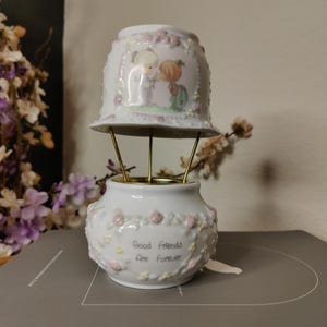 May include: A white ceramic Precious Moments figurine with a bell-shaped top and a vase-like base, connected by gold-colored metal rods. The top features an illustration of two children. The base has the text "Good Friends Are Forever" and is decorated with pink flowers.
