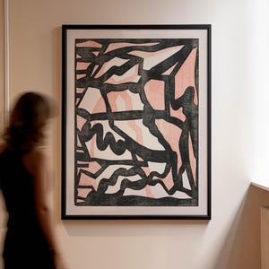 May include: Abstract art print in a black frame. The artwork features a design with black, white, and pink shapes. The print is displayed on a light-coloured wall. A person in a black dress is slightly blurred in the foreground.