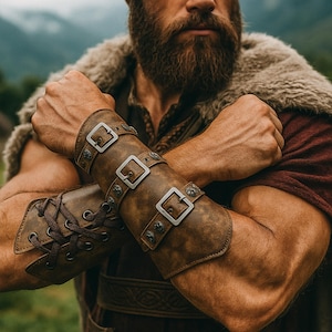 Peut inclure: Brassards en cuir marron avec boucles argentées et clous décoratifs. Les brassards sont lacés sur le côté et portés sur une chemise rouge. Une cape doublée de fourrure est drapée sur les épaules.