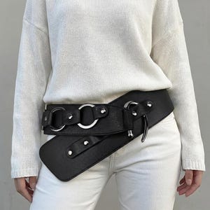 May include: Black faux leather belt with silver-tone metal rings and rivets. The belt features an asymmetrical design with a wide front panel and a wrap-around style, creating a unique and stylish accessory.