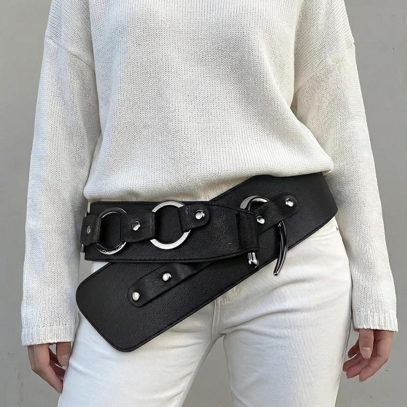 May include: Black faux leather belt with silver-tone metal rings and rivets. The belt features an asymmetrical design with a wide front panel and a wrap-around style, creating a unique and stylish accessory.