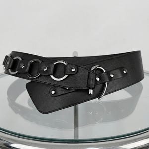 May include: A black belt with silver-tone metal rings and rivets. The belt features a wide design with an overlapping closure and a decorative metal ring detail. The belt is made of a smooth, faux leather material.