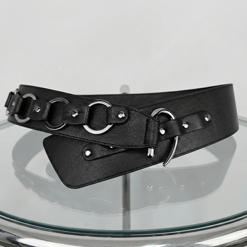 May include: A black belt with silver-tone metal rings and rivets. The belt features a wide design with an overlapping closure and a decorative metal ring detail. The belt is made of a smooth, faux leather material.