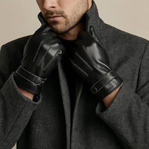 以下が含まれることがあります： 白いステッチとボタン留めのリストストラップが付いた黒い革手袋。手袋はグレーのコートと一緒に着用されています。手袋は柔らかい革素材で作られているようです。