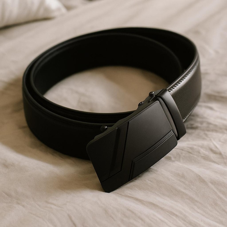 Op de afbeelding: Een zwarte riem met een matte afwerking en een rechthoekige gesp. De gesp heeft een geometrisch ontwerp. De riem is opgerold op een neutraal gekleurd oppervlak.