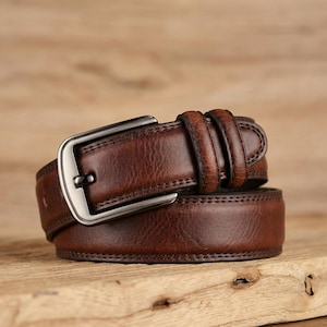 Op de afbeelding: Een bruine leren riem met een zilverkleurige gesp. De riem is gemaakt van getextureerd leer en heeft een klassiek ontwerp. De gesp is rechthoekig en heeft een enkele pin. De riem is ongeveer 3,8 cm breed.