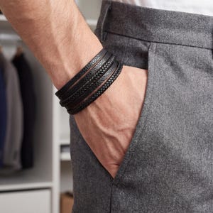 Op de afbeelding: Een stapel zwarte leren armbanden met verschillende texturen, waaronder gevlochten en gladde ontwerpen. De armbanden worden om een pols gedragen, contrasterend met de grijze stof van de broek. De afbeelding suggereert een focus op accessoires.