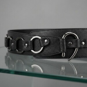 May include: A black belt with silver-colored metal rings and a hook closure. The belt is made of a textured material and features multiple decorative rings along its length. The hook closure is a large, curved design.