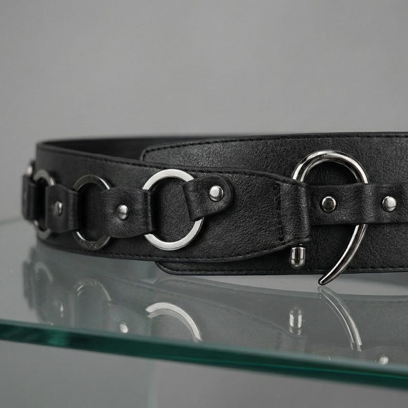 May include: A black belt with silver-colored metal rings and a hook closure. The belt is made of a textured material and features multiple decorative rings along its length. The hook closure is a large, curved design.