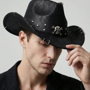 Könnte beinhalten: Schwarzer Cowboyhut mit strukturierter Oberfläche, verziert mit silbernen Nieten und einem Totenkopf-Emblem auf dem Band. Der Hut hat Metallringe am Rand und bietet eine einzigartige, kantige Ästhetik.