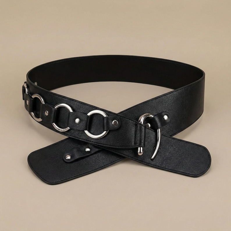 May include: Black belt with silver-tone metal ring and hook closure. The belt is made of a smooth material and features a series of decorative rings. The belt is designed to be worn around the waist.
