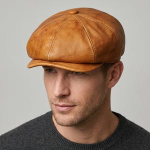 Puede incluir: Gorra de estilo newsboy de cuero marrón claro. Presenta una corona redondeada, una pequeña visera rígida y costuras visibles. Un botón adorna la parte superior.