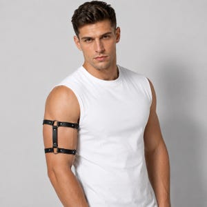 May include: A black leather arm harness featuring two horizontal straps connected by a vertical center strip, secured with silver-toned metal rings and rivets. The accessory is worn around the upper arm, providing a structured, adjustable fit.