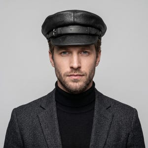 May include: A black leather cap with a rounded crown and a short, stiff brim. The cap features a leather band with a buckle detail. The person in the image is wearing a black turtleneck and a gray blazer.