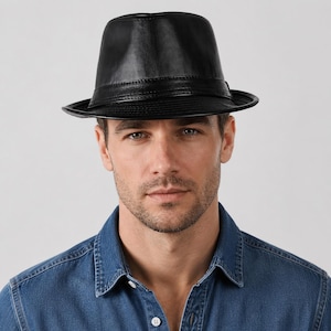 Puede incluir: Un sombrero fedora negro con un acabado liso y una banda negra a juego alrededor de la base de la copa. El sombrero presenta un ala estructurada que se curva ligeramente hacia arriba en los lados y una copa pellizcada, ofreciendo una silueta clásica.