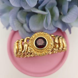 May include: A gold-toned bracelet with a large, round, red gemstone at its center. The bracelet features an ornate, detailed design around the gemstone and on the connecting links. The bracelet is displayed on a pink surface with blue flowers in the background.
